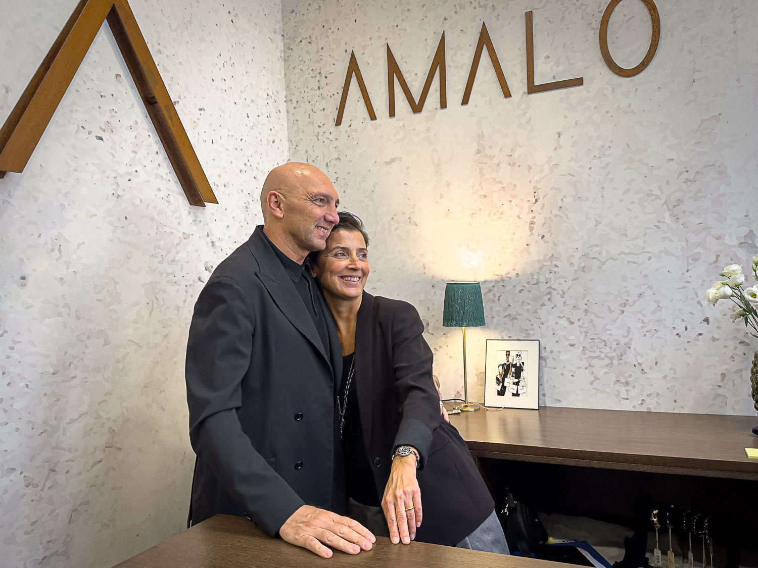 Angela e Luciano abbracciati al bancone in legno sotto la scritta AMALO, parete chiara effetto terrazzo e lampada verde